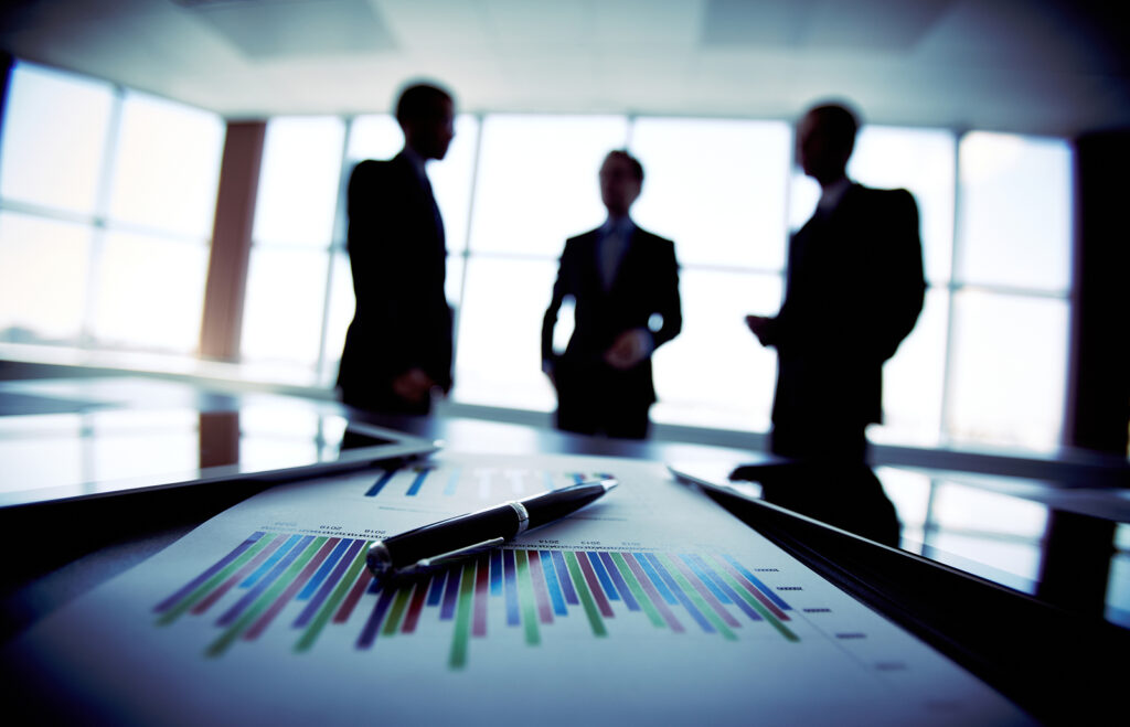 Em primeiro plano uma caneta e gráficos empressos. em segundo plano três pessoas conversando.